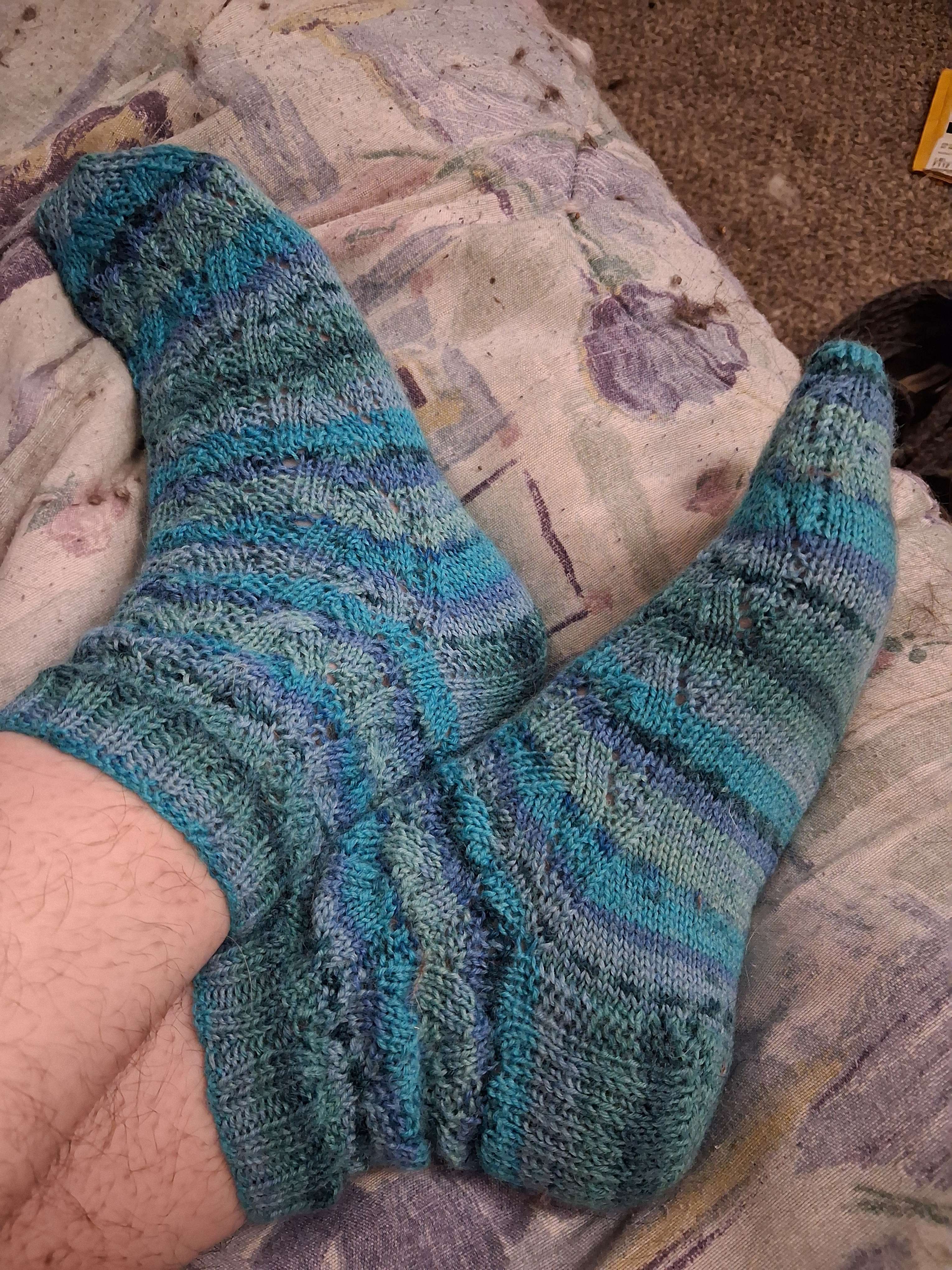 A pair of knitted socks made of varigated blue yarn are worn against the background of a crumpled duvet on a bed.